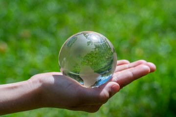 Glass globe in girl hand on blurred green grass banner background with sunlight in city. Concept of environment world earth day. Protect clean nature and ecology, sustainable lifestyle, save planet