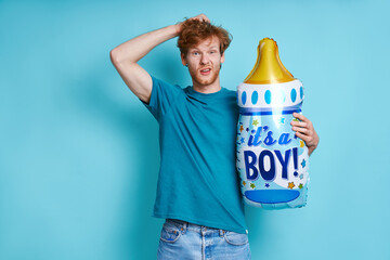 Surprised redhead man holding baby bottle shaped balloon while standing against blue background