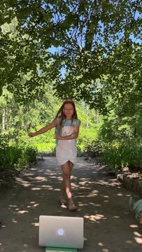 a little energetic girl dances in a park in nature in front of a laptop in summer, a girl actively spends time outdoors, dancing and having fun, a girl with long hair shows or watches a dance master c