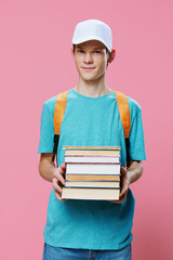 a handsome, funny guy in a blue t-shirt and a cap on his head stands holding textbooks in his hands with a backpack on his back