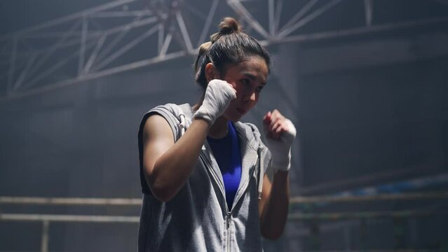 4K Strong Asian sportswoman athlete do combat sport training workout boxing exercise with punching bag in abandoned building. Active woman boxer practicing fighting exercise kickboxing in dark old gym