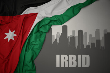 abstract silhouette of the city with text Irbid near waving national flag of jordan on a gray...