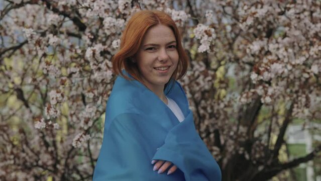 Beautiful redhead woman covering shoulders with Ukrainian flag in spring cherry garden. Smiling caucasian female looking at camera and calling for support Ukraine during war.