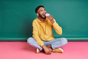 Cheerful African man enjoying doughnut while sitting against colorful background