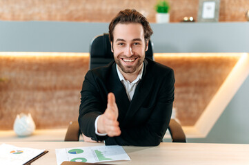 Positive successful confident caucasian businessman in a suit, sitting at a desk in the office, holding out his hand for a handshake, making a deal, agreement, contract, cooperation, smiling friendly