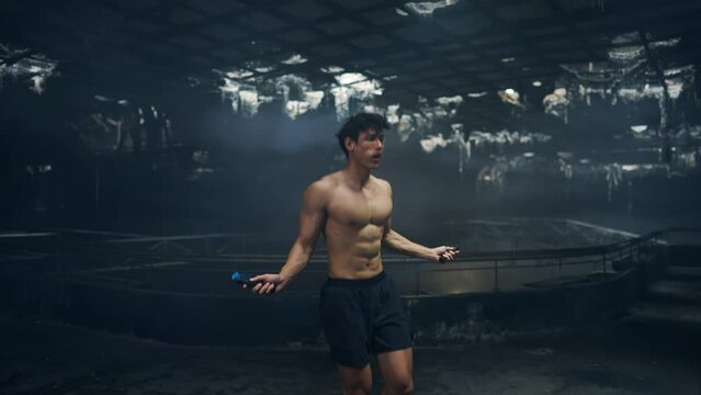 Strong Asian sportsman do combat sport training cardio workout skipping jump rope exercise in abandoned building. Shirtless active man bodybuilder practicing muscular strength training in dark old gym