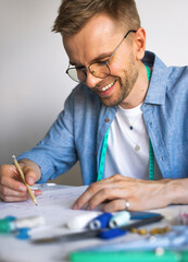 A DIY designer draws a sketch for a new costume project. A seamster with glasses is working at the table. A smiling man in a blue shirt makes a sketch for sewing clothes. Real emotions, life style