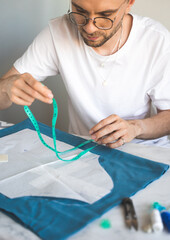 A self-taught seamster in white t-shirt and glasses works with fabric, pattern and chalk. A tailor uses a measuring tape during his work in a home workshop. Small business, life style concept