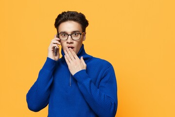 horizontal photo of a cute, surprised man standing on an orange background in a blue zip-up jacket and black eyeglasses, talking on the phone with his hand covering his mouth.