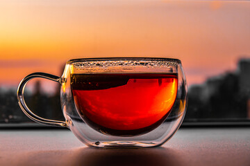 Close up of tea with lemon in transparent cup on table and city at sunset on background. Hot drink...