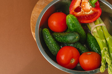 Vegetables lie in a metal bowl: tomatoes, asparagus, cucumbers, red bell peppers. on a wooden board and brown background. place for text. view from above