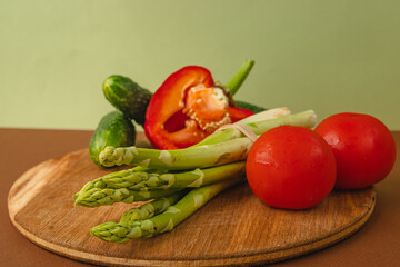 Vegetables lie on a wooden board: tomatoes, asparagus, cucumbers, red bell peppers. brown, light green background. place for text.