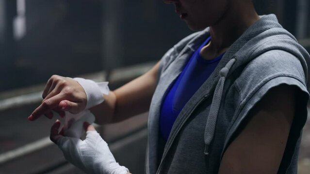 4K Strong Asian woman athlete wraps her hands with boxing bandages while do sport training workout boxing in abandoned building. Female boxer practicing fighting exercise kickboxing in dark old gym