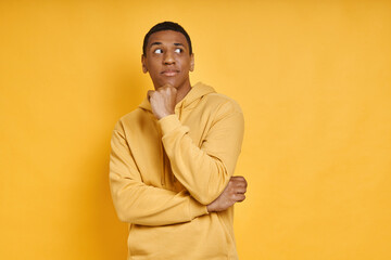 Thoughtful African man holding hand on chin while standing against yellow background