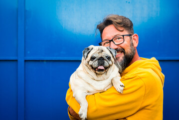 Portrait of man and dog. Young adult handsome with beard and eyewear hug and hold funny nice pug both looking on camera and blue background wall. Copy space and color. Love and friendship