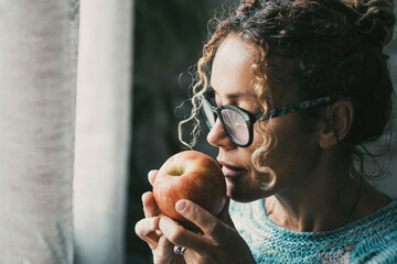 Side portrait of woman holding an apple and looking outside the window at home. Concept of healthy...