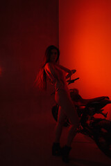 young teen brunette girl biker is standing fashion like silhouette in profile and smiling near black sports motorcycle on red studio wall background in backlight. lifestyle fashion concept, free space
