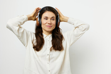funny woman in light shirt stands listening to music in her headphones, closing her eyes with pleasure and holding headphones with her hands. Horizontal photo on a light background with an empty space