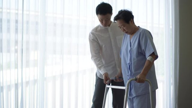 Adult Asian son help mother do physical therapy using walker in hospital love encourage and care family