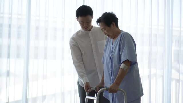 Adult Asian son help mother recovery therapy using walker to exercise in hospital loving caring huging