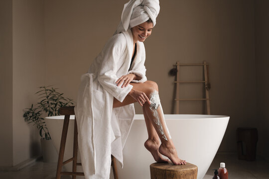 Woman depilating legs using shaving foam and razor, dressed in bathrobe standing in bathroom