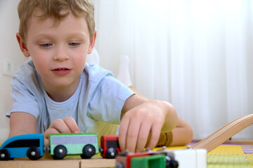 Close-up of wooden toys on puzzle mats that the boy is playing with. A child lying on the floor rolls a train with colorful cars along a wooden railway.