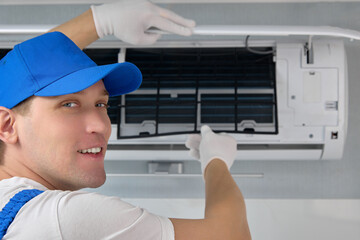 Professional repairman of air conditioning equipment replaces the filter elements. Smiling man in uniform and gloves removes the grilles from the air conditioner for cleaning and prevention