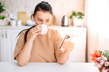 A beautiful woman drinks coffee and looks at the phone in the kitchen. A young woman enjoys the morning.