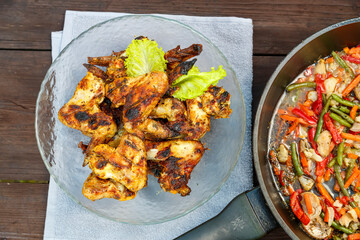 A frying pan with stewed vegetables on a wooden table next to a dish with grilled wings.