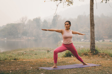 Fototapeta na wymiar Asian woman practicing yoga in Warrior Pose (Virabhadrasana) on the mat beside the tree and lake in outdoor park.