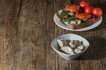 On a wooden background there are plates with ready-made cod liver and ready-made healthy sandwiches with tomatoes and herbs.