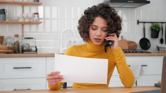 Businesswoman talking on mobile phone with financial report in hand at home kitchen. Hispanic curly woman working communicates with employee using smartphone, sitting on chair at desk. Laptop computer