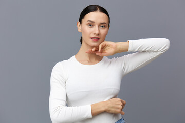 a beautiful, slender, elegant woman stands on a gray background in a white T-shirt with long sleeves and gently touches her neck with her fingers while looking at the camera, smiling pleasantly.