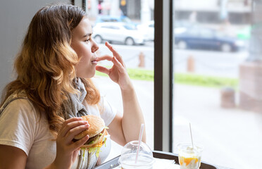 Asian young girl sitting at table indoors, holding hamburger in her hand, looking out window and from pleasure licking her fingers. Concept: fatty, high-calorie, unhealthy food. .