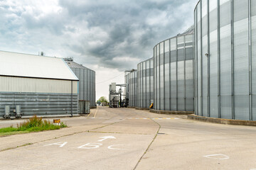 Fototapeta na wymiar Dramatic view of steel grain silos seen against a looming storm. The concrete roadway denotes the route for HGVs to unload the grain to the silos.