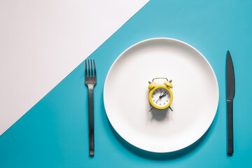 Alarm clock on a white plate with a knife and fork on blue background.