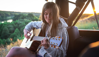 A young woman plays the acoustic guitar in the trunk of a car in nature.