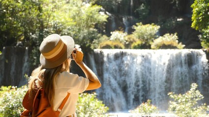 Female travel blogger with photo camera shooting amazing tropical waterfall in Asia journey. Cinematic and inspiration video of photographer woman taking picture of nature in travel vacation. - obrazy, fototapety, plakaty