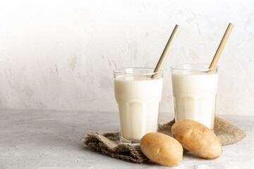 Two glasses of vegan potato milk with bamboo drinking straws and potato tubers on neutral backdrop.