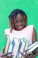 A smiling African-American woman holds a stack of books on a green background. Portrait. Back to...