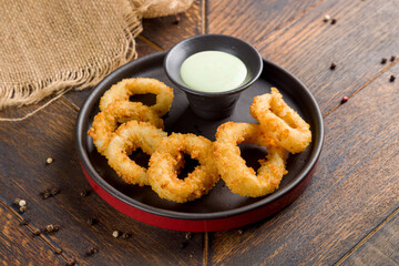 Rings of squid in batter with sauce on dark plate on wooden table