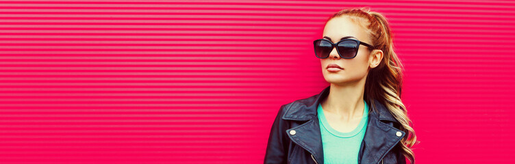 Portrait of beautiful blonde young woman wearing sunglasses, black rock jacket on pink background