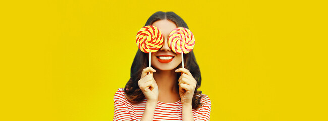 Portrait of cheerful smiling young woman covering her eyes with lollipop on yellow background