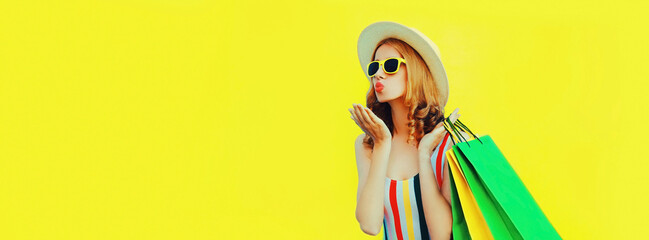 Portrait of beautiful happy young woman blowing her lips sending air kiss with shopping bags wearing summer straw hat on yellow background