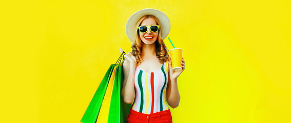 Portrait of happy smiling young woman drinking fresh juice with colorful shopping bags wearing summer straw hat on yellow background