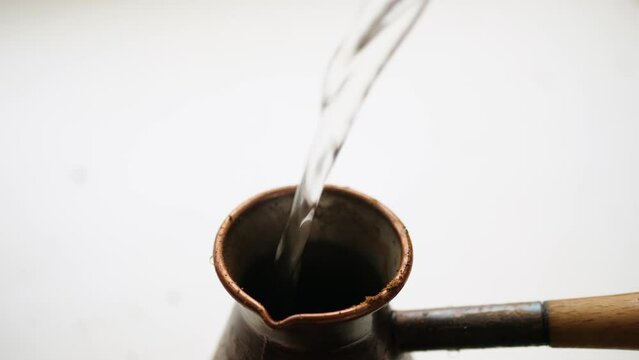 Close up boiling water pours into the brown copper cezve with freshly ground coffee on white countertop, soft focus with copy space