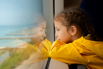 A little girl of 6-7 years old looks out the window of a train at the sea. She's wearing a yellow parka. Journey. Reflection. Vacation. Summer. Family vacation.