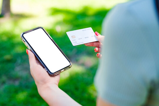 Young caucasian woman wearing tee standing on city park, outdoors holding blank screen mobile phone credit bank card do online shopping provide internet payment. Safe mobile banking.