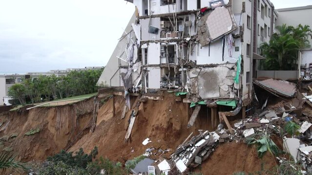 Pan from left to the destruction of an apartment building that has collapsed into a landslide caused by severe storm damage. Two cars are balanced on the edge of a collapsed driveway.