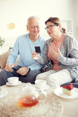 Elderly attractive lady in eyeglasses sitting on sofa and gesturing hand while sharing story of past with husband and showing old photographs to him